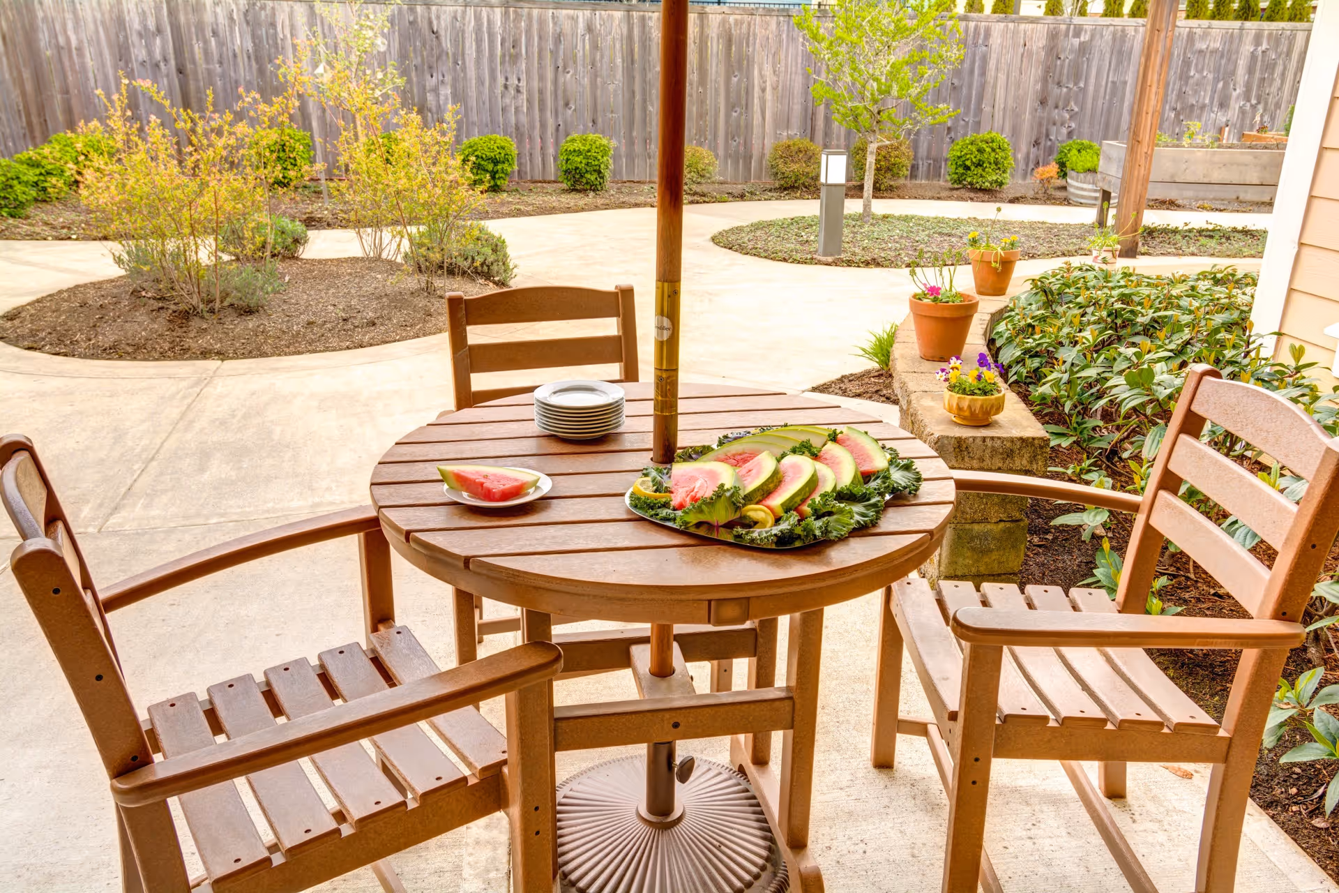 Outdoor patio area with a round wooden table and three wooden chairs. On the table, there is a plate with sliced watermelon and a stack of small plates. The patio is surrounded by a garden with various plants and a wooden fence in the background.