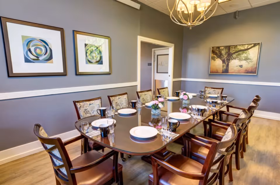 A formal dining room with a long polished wooden table set for eight people. The table has white plates, black mugs, glasses, and small floral centerpieces. The room has gray walls adorned with framed artwork and a chandelier hanging from the ceiling. There is a door in the background.