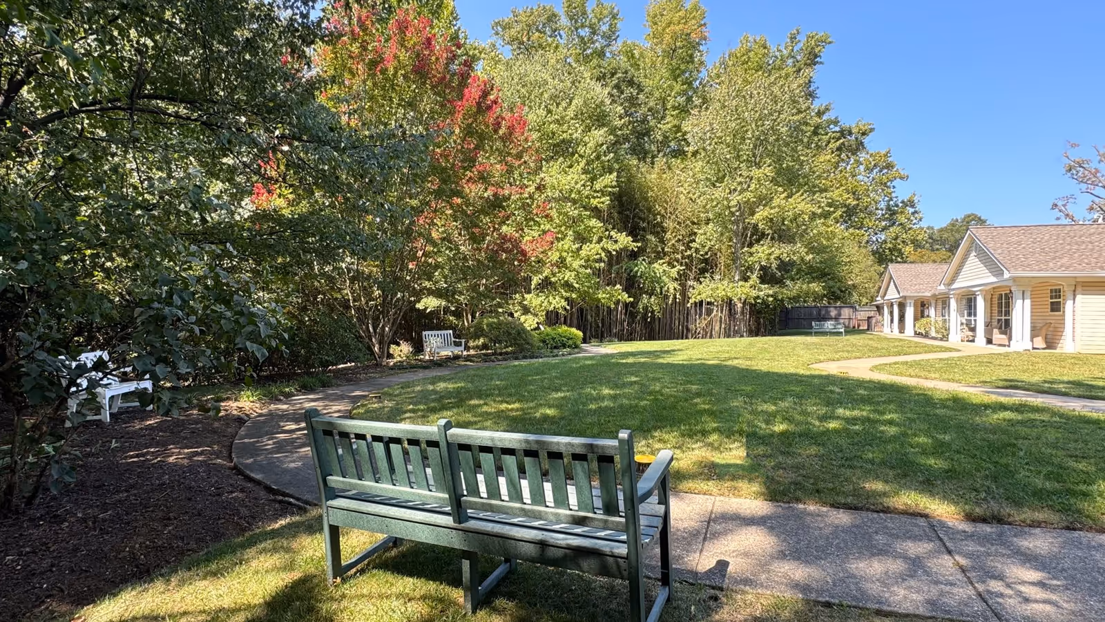 A peaceful outdoor garden area at Charter Senior Living of Annapolis featuring green grass, several benches, a curved concrete pathway, and trees with some leaves turning red under a clear blue sky.