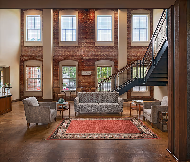 A spacious living area with exposed brick walls and large windows letting in natural light. The room features a patterned sofa, two armchairs, a red area rug, and small wooden tables. A modern black metal staircase with a railing leads to an upper floor. The floor is wooden, and there are additional chairs and a small table near the windows.