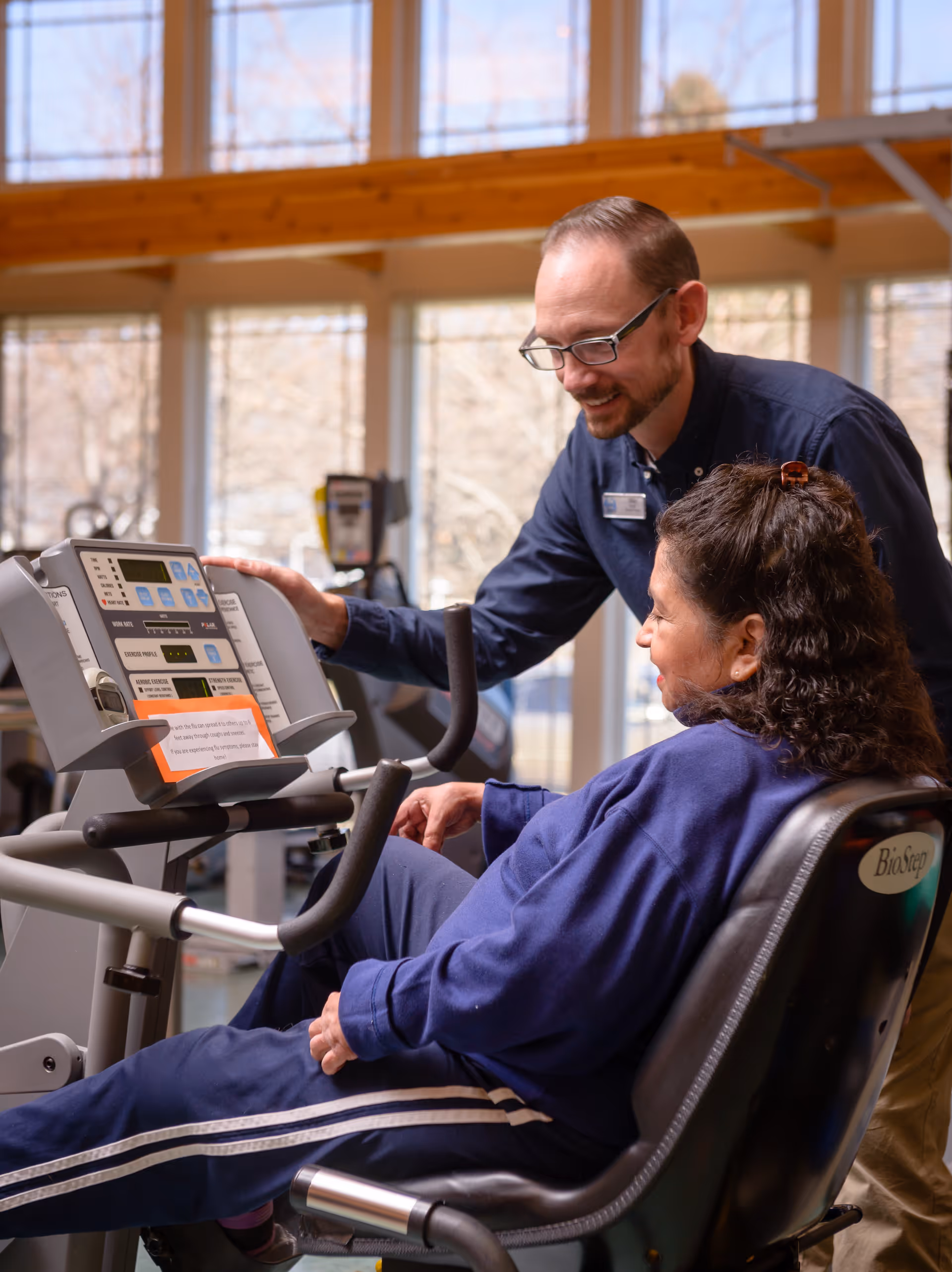 A man wearing glasses and a name tag is assisting a woman seated on a recumbent exercise bike in a bright room with large windows. The woman is dressed in a blue tracksuit and appears to be receiving guidance on using the exercise equipment.