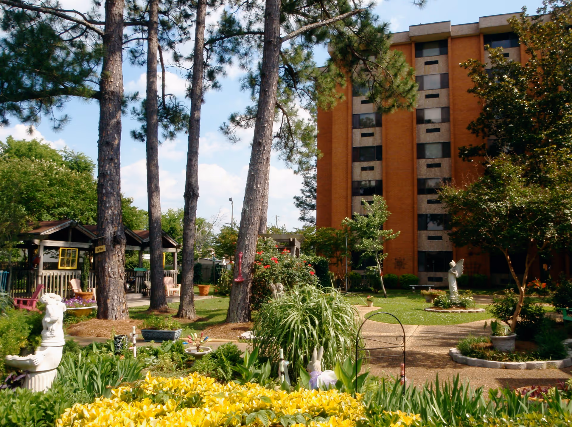 A vibrant garden area with yellow flowers, green plants, and decorative statues in front of a multi-story brick building. Tall pine trees and a wooden gazebo with chairs are visible, along with a clear blue sky.