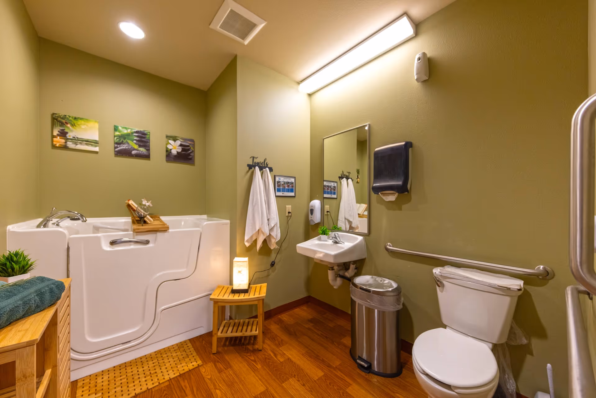 A bathroom with a walk-in bathtub, a small sink with a mirror above it, a toilet with a grab bar, a trash can, and a wooden stool with a lit lamp. The walls are painted green and decorated with three small nature-themed pictures. There are towels hanging on a hook and a paper towel dispenser mounted on the wall.