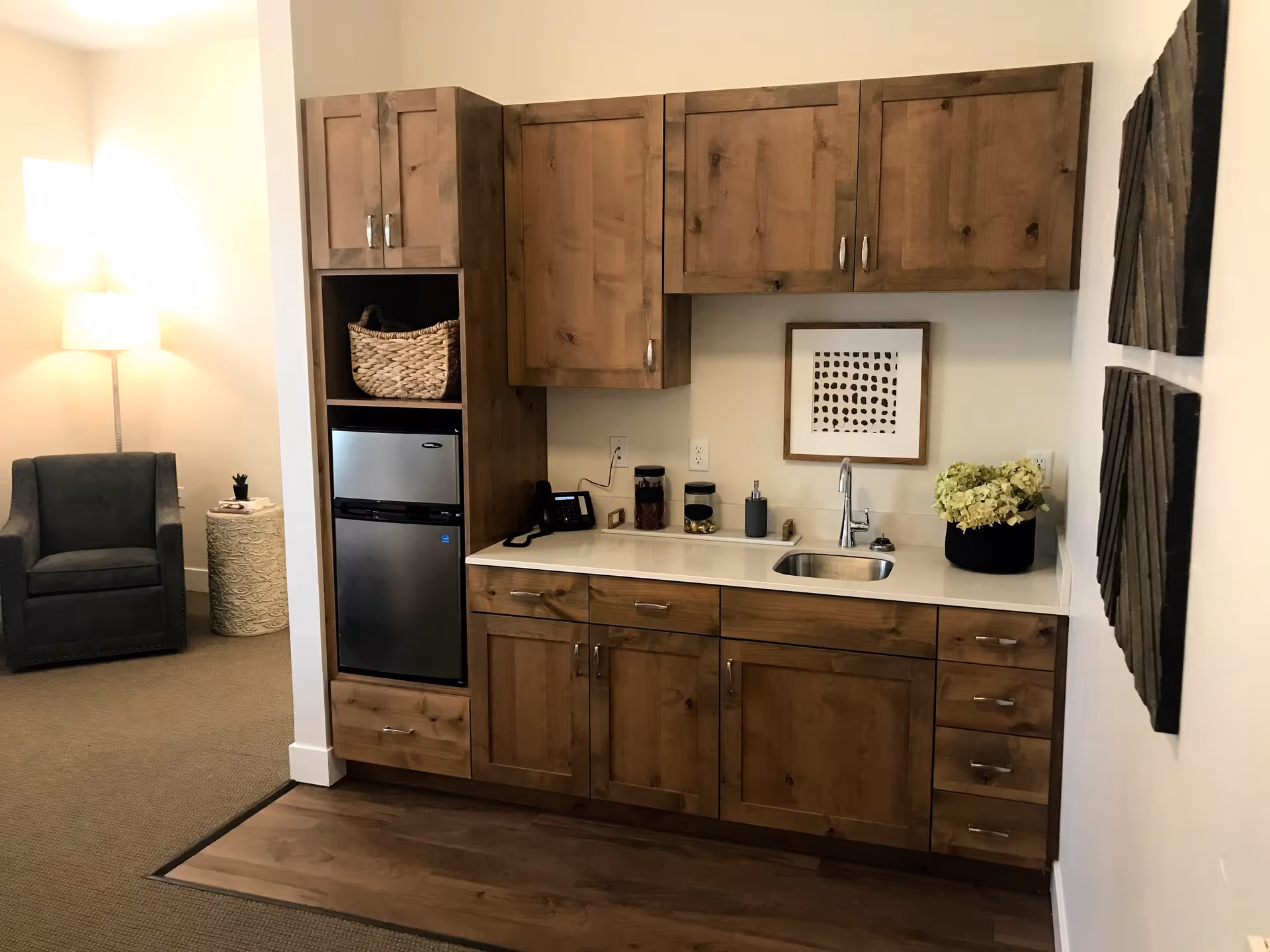 A small kitchenette area with wooden cabinets, a mini refrigerator, a sink, and countertop with decorative items including a framed abstract artwork and a potted plant. To the left, there is a gray armchair, a round side table, and a floor lamp in a softly lit room.
