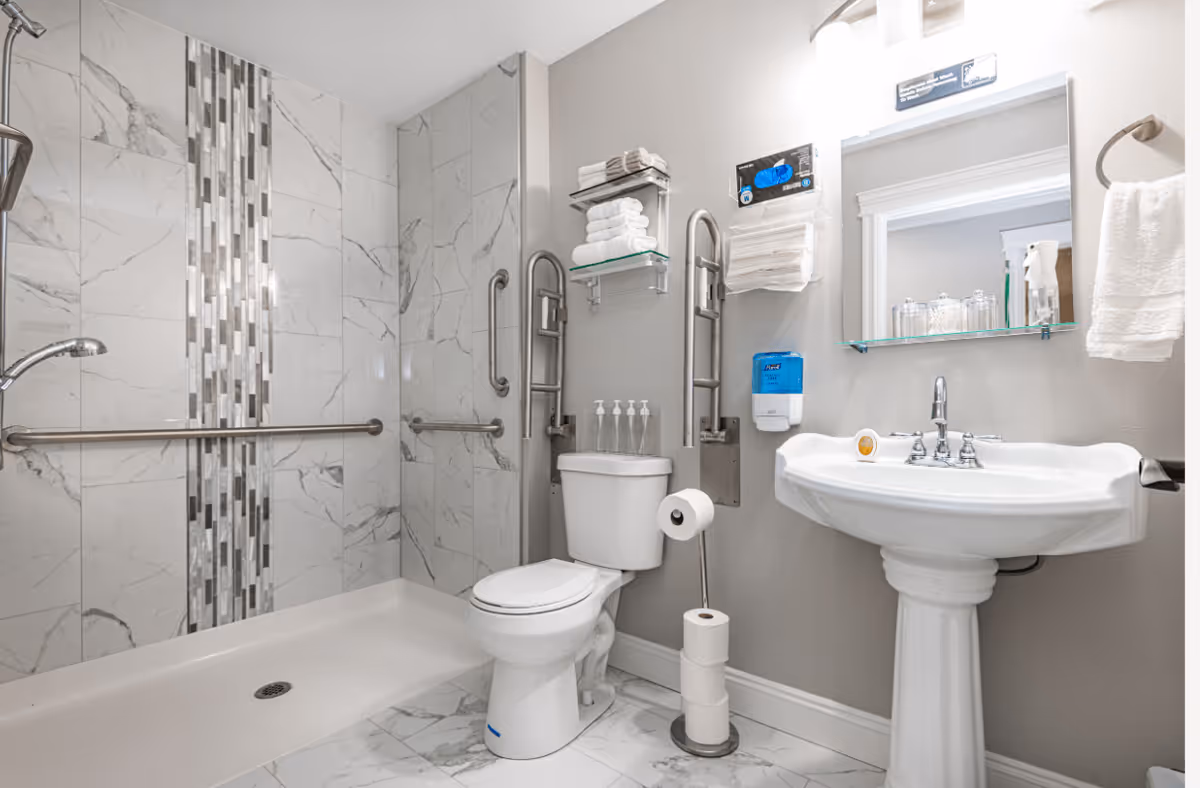 A clean and modern bathroom with a walk-in shower featuring marble-patterned tiles and vertical mosaic accent strip. The bathroom includes a white toilet with safety grab bars on the walls, a pedestal sink with a mirror above it, and shelves holding neatly folded towels and toiletries. There is a wall-mounted soap dispenser and a stand holding extra toilet paper rolls.