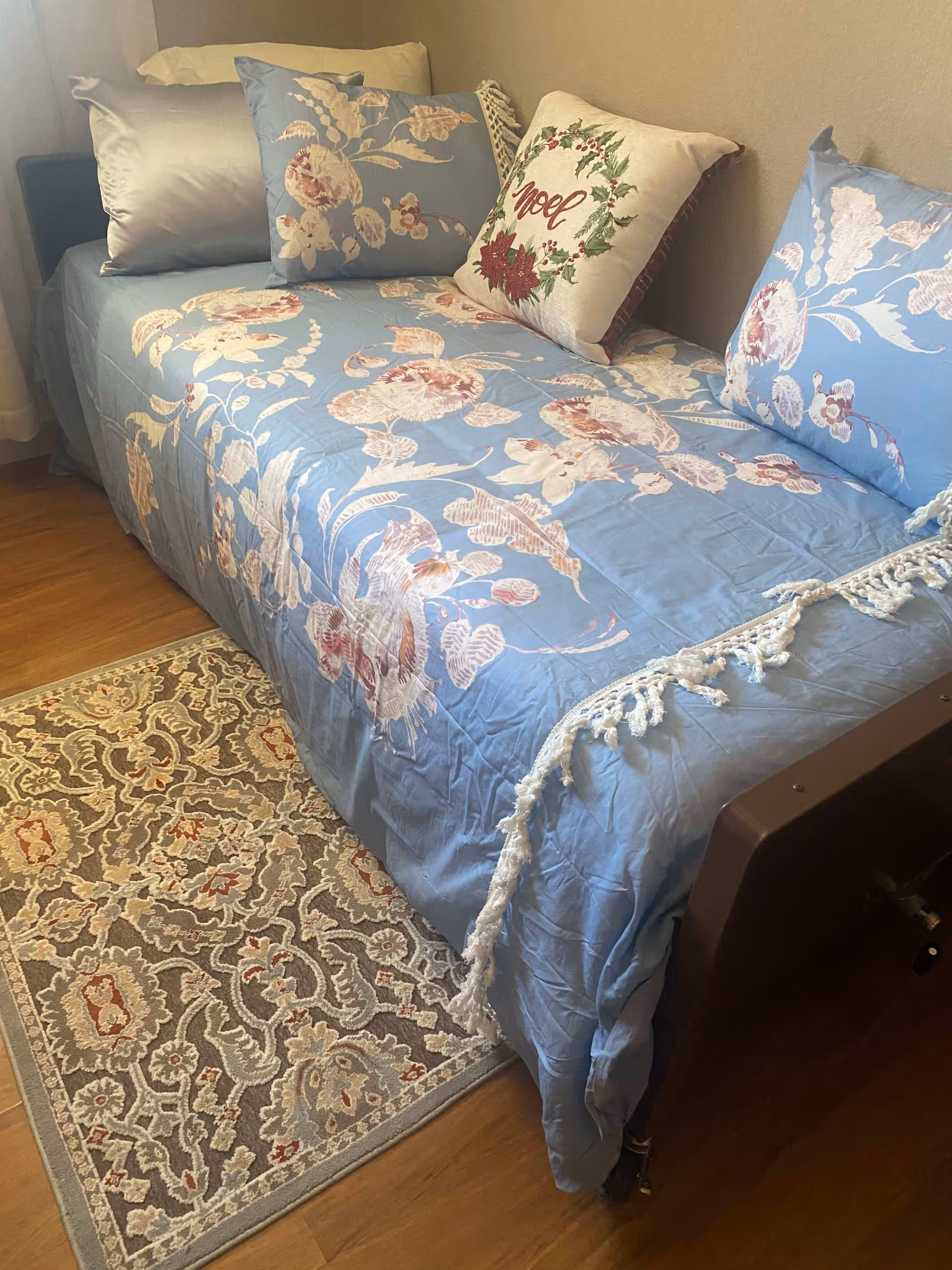 A single bed with a blue floral bedspread and matching pillows, including one decorative pillow with the word 'Noel' and a holiday wreath design. The bed is placed against a beige wall and next to a window with white curtains. A patterned area rug is on the wooden floor beside the bed.