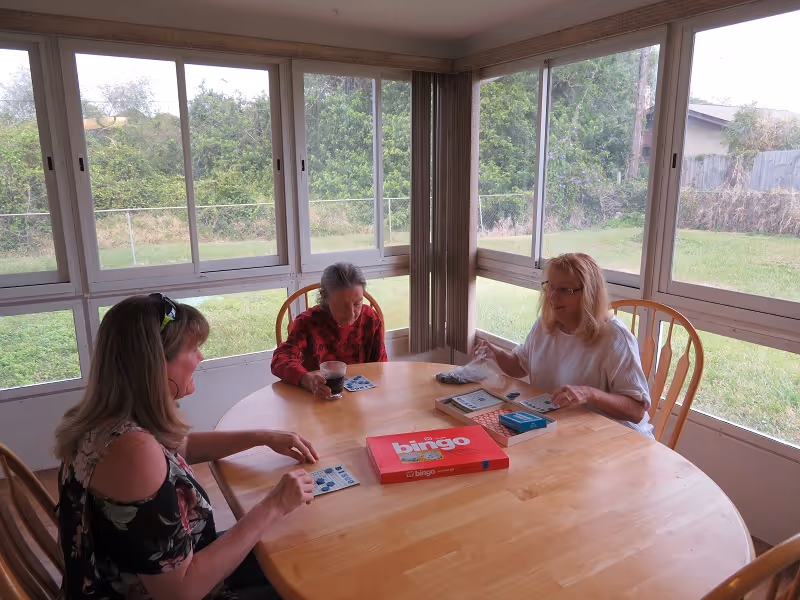 Three women sitting around a wooden table playing bingo in a sunroom with large windows overlooking a grassy yard.