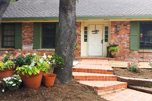 Front entrance of a brick house with a white door, green shutters, brick steps and potted flowering plants.