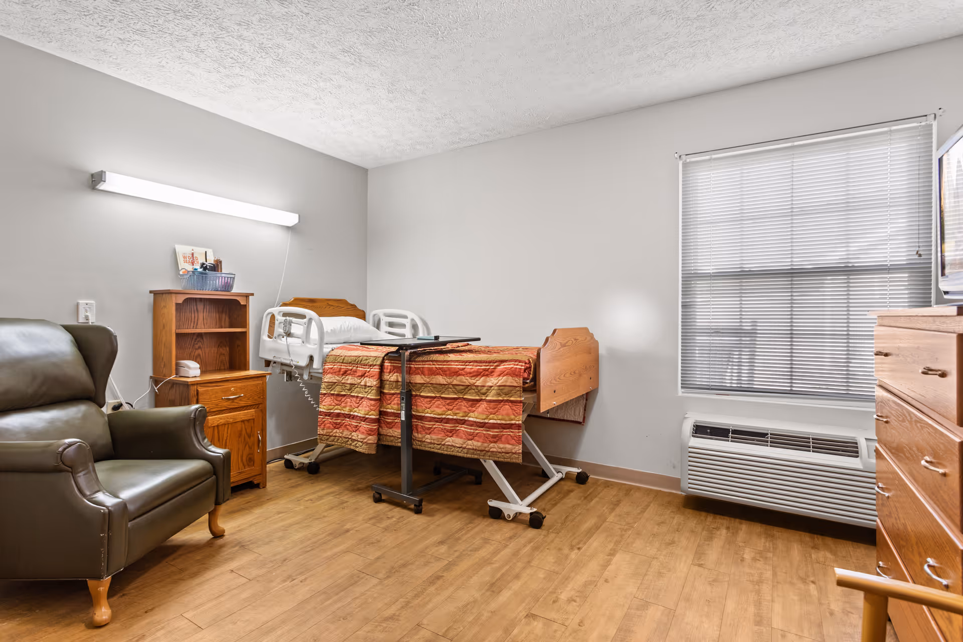 A senior living facility bedroom with a hospital-style bed covered with a red and gold striped blanket, a wooden nightstand with shelves and a drawer, a green leather armchair, a wooden dresser, a window with blinds, and a wall-mounted light fixture.