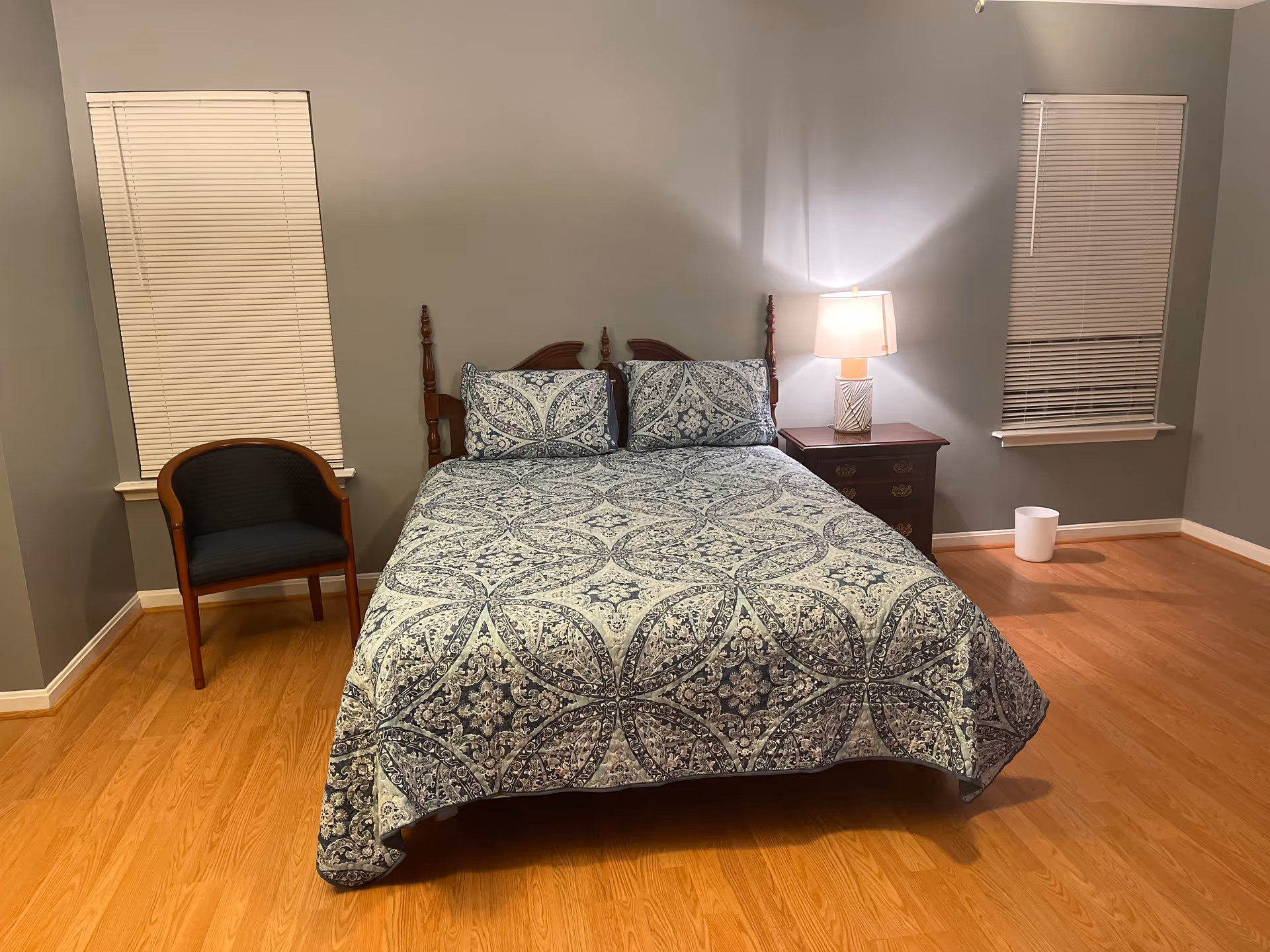 A bedroom with a wooden bed frame and a bed covered with a blue and white patterned bedspread and matching pillows. There is a wooden nightstand with a lamp on the right side of the bed, a wooden chair with a dark cushion on the left side near a window with closed blinds, and another window with closed blinds on the right side. The floor is wooden, and there is a small white trash bin near the right window.