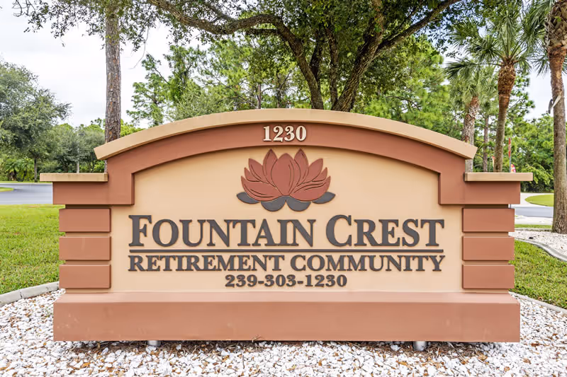 A large outdoor sign for Fountain Crest Retirement Community with the address number 1230 displayed at the top. The sign features a decorative lotus flower design and the phone number 239-303-1230 at the bottom. The sign is set in a landscaped area with white rocks and green grass, surrounded by trees and a paved road in the background.
