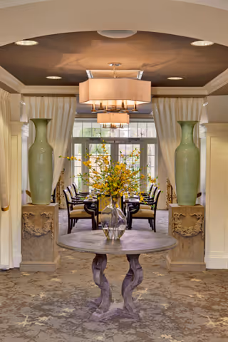 Elegant interior space featuring a round wooden table with a glass vase holding yellow flowers in the foreground. Behind the table, two large green vases are placed on stone pedestals on either side of an entrance framed by cream-colored curtains. Through the entrance, a dining area with a table and chairs is visible, illuminated by modern ceiling lights.