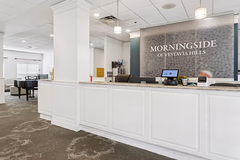 Reception area of Morningside of Vestavia Hills featuring a long white front desk with a computer and office supplies. Behind the desk is a decorative wall with the facility's name displayed. The space is well-lit with ceiling lights and has a carpeted floor. In the background, there is a seating area with a grand piano and windows with blinds.