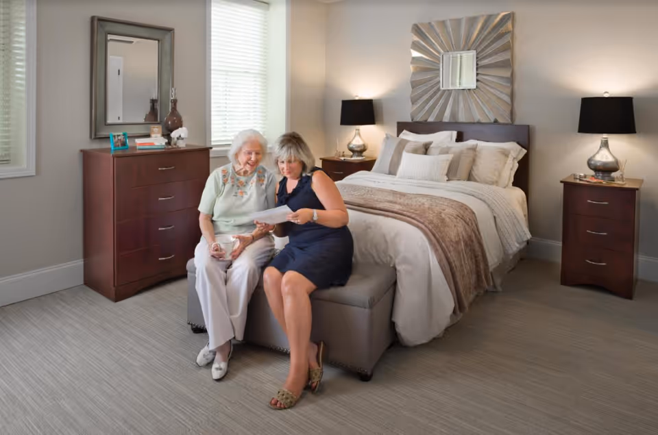 An elderly woman and a younger woman sitting on a bench at the foot of a neatly made bed in a well-lit bedroom. The elderly woman is holding a cup while the younger woman shows her a document. The room features two wooden dressers with lamps, a decorative mirror above the bed, and windows with blinds.