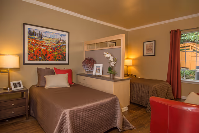 A cozy bedroom with two single beds separated by a wooden divider. Each bed has brown bedding and decorative pillows. There are two bedside tables with lamps, a framed colorful landscape painting on the wall, a window with red curtains, and a red armchair in the foreground.