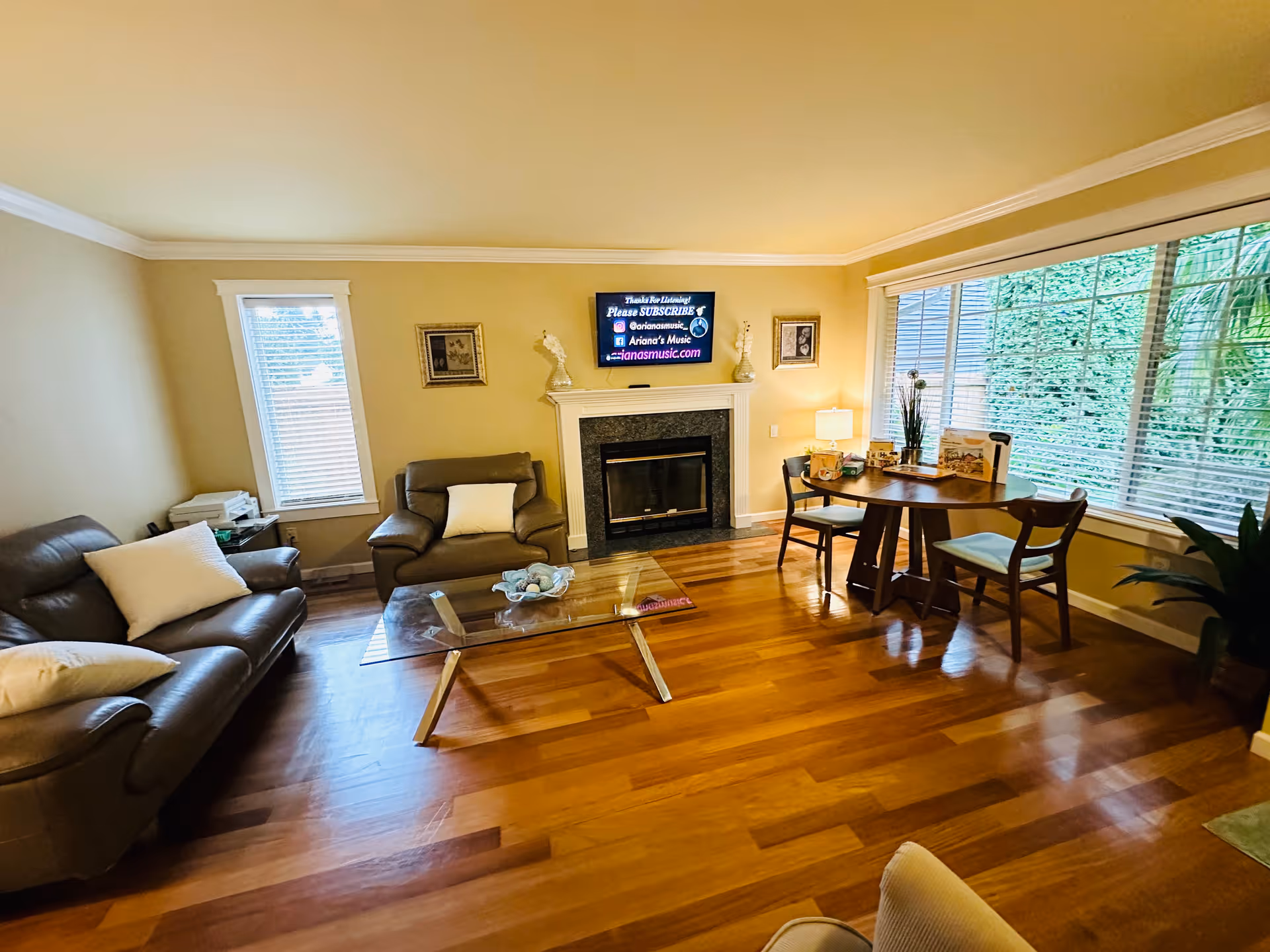 A cozy living room with wooden flooring, a glass coffee table, two brown leather sofas with white pillows, a fireplace with a TV mounted above it, a round wooden dining table with four chairs near a large window with blinds, and decorative plants and lamps.