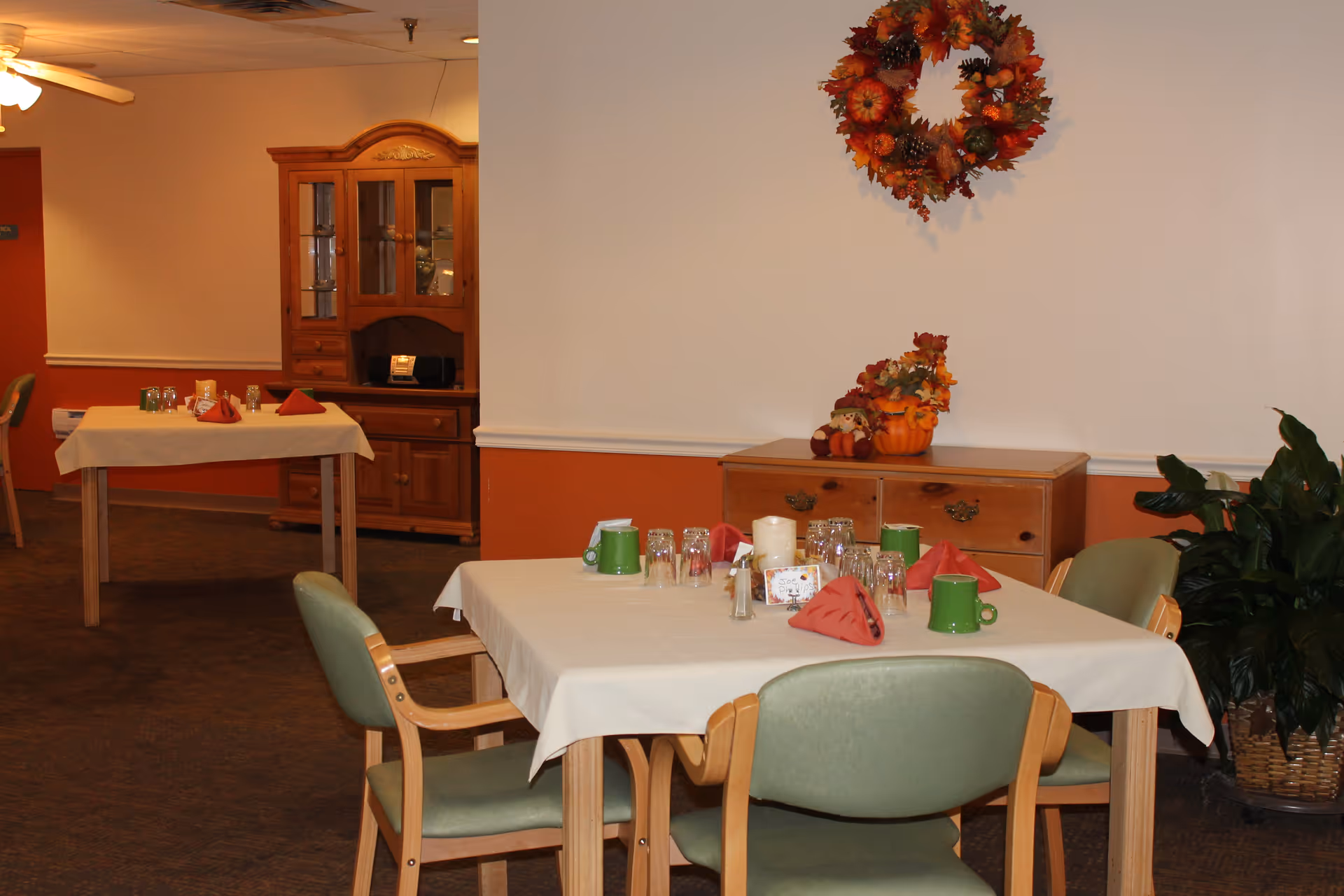 A dining room with tables set with cups, napkins and glassware, green chairs, and fall decorations on a sideboard and wall.