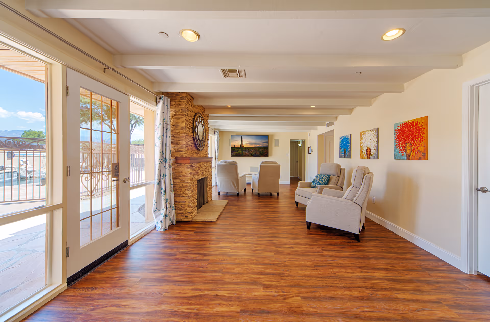 A bright and spacious living room with wooden flooring, beige walls, and white ceiling beams. The room features a stone fireplace with a large clock above it, several beige armchairs arranged facing a wall-mounted flat-screen TV, and colorful artwork on the wall. Large glass doors and windows on the left side provide a view of an outdoor area with a pool and trees.