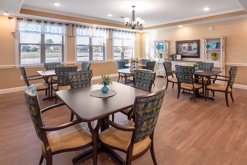 A bright dining room with several square tables, each surrounded by four cushioned chairs with patterned backs and tan seats. The room has large windows with sheer curtains allowing natural light to fill the space. A chandelier hangs from the ceiling, and a white sideboard with decorative items and a framed painting is against the far wall. The floor is wood, and the walls are painted beige with white trim.