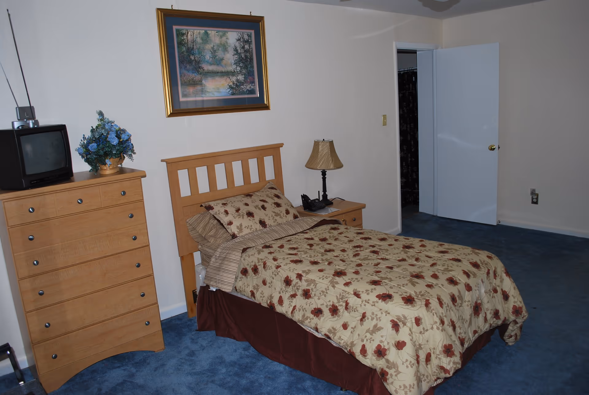 A bedroom with a single bed covered in a floral patterned bedspread. Next to the bed is a wooden nightstand with a lamp and a telephone. To the left of the bed is a wooden dresser with a small TV and a vase of blue flowers on top. A framed landscape painting hangs on the wall above the bed. The room has blue carpet and white walls, with an open door leading to another room.