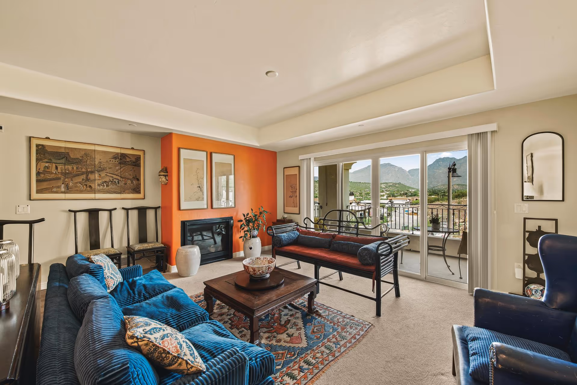 A cozy living room with a blue velvet sofa, a wooden coffee table on a patterned rug, and a black metal bench with red cushions. The room features an orange accent wall with a fireplace, framed artwork, and a large sliding glass door leading to a balcony with a view of mountains in the background.