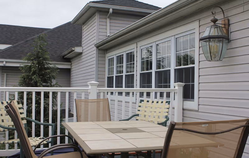 Patio deck with a tiled table and several chairs in front of a beige-sided building with multiple windows.