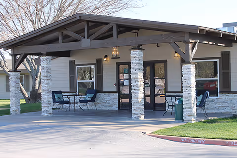 Front exterior view of a single-story building with a covered entrance supported by stone pillars. There are outdoor chairs and tables on the porch area, and a tree is visible in the background.