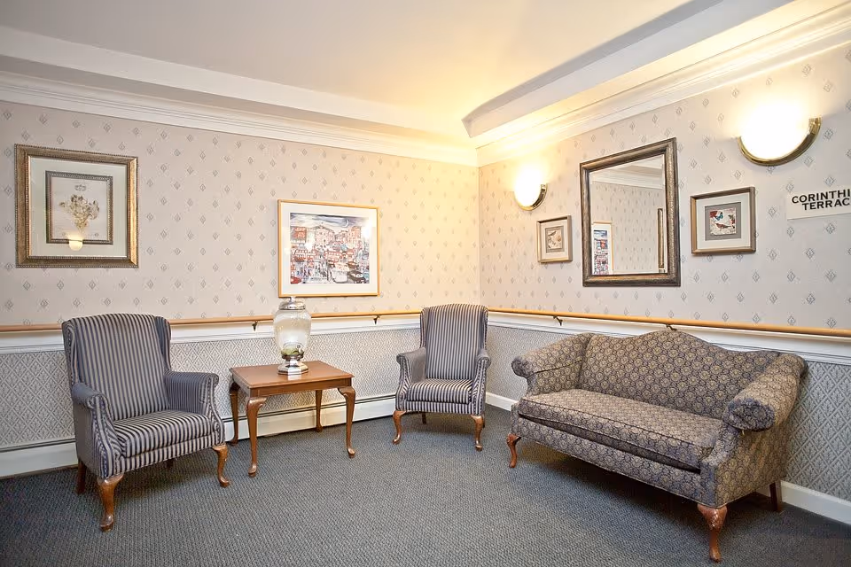 A cozy sitting area with two striped armchairs and a patterned sofa arranged around a wooden side table with a decorative glass jar. The walls are decorated with framed artwork and a large mirror, with soft wall sconces providing warm lighting.