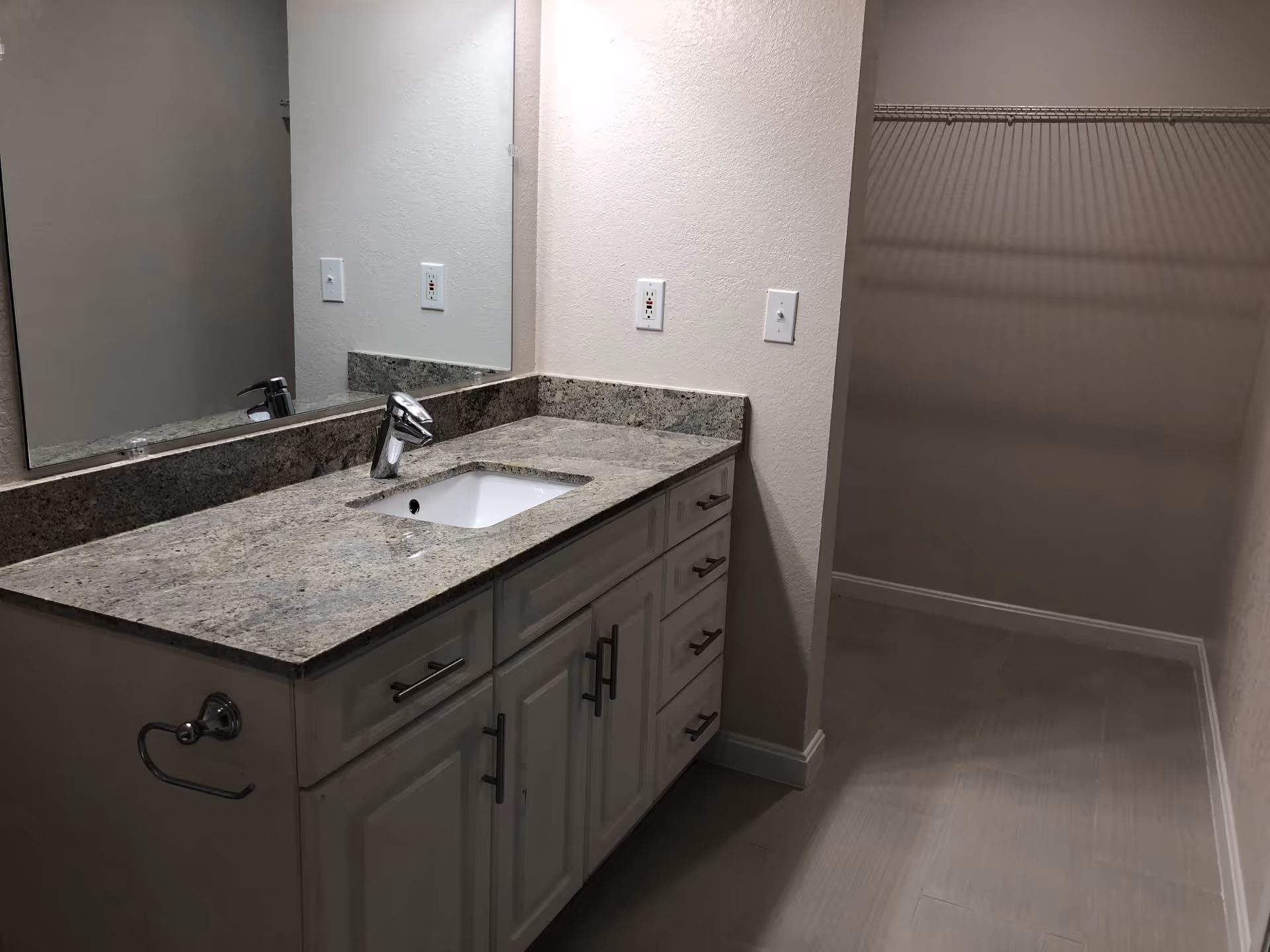 A bathroom vanity with a granite countertop, a built-in sink, and a modern faucet. There is a large mirror above the vanity and a towel ring mounted on the side. To the right, there is an open walk-in closet with a wire shelf for hanging clothes. The floor is tiled and the walls are painted light beige.