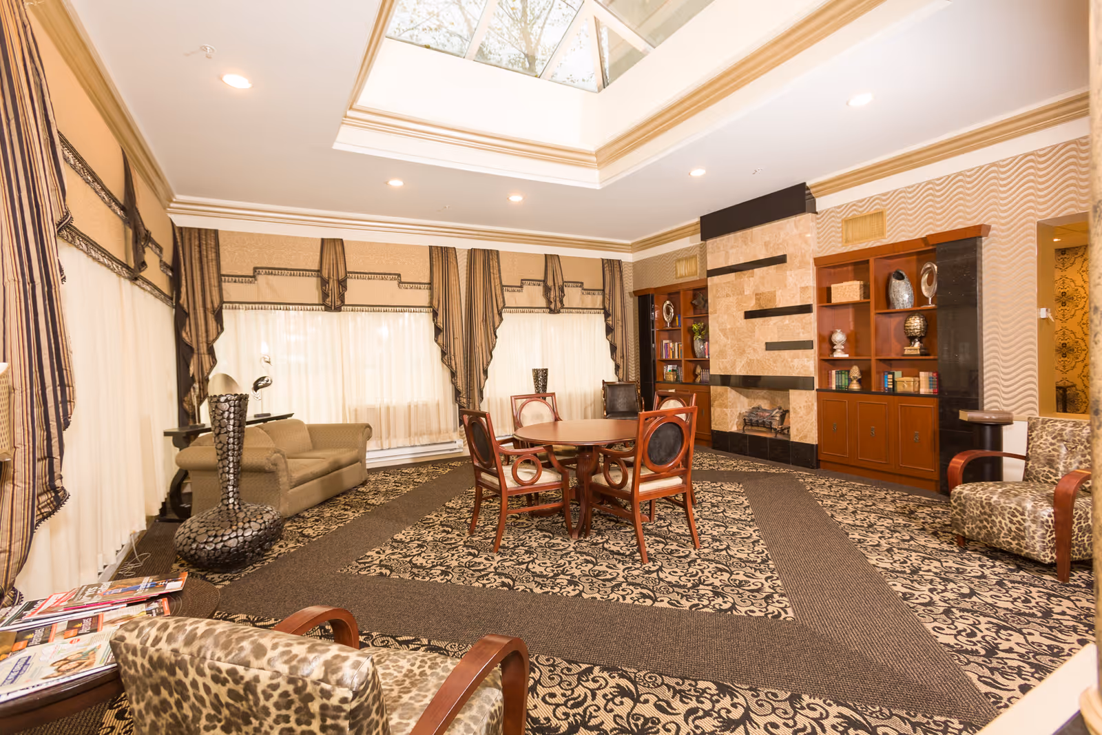 A spacious and elegantly decorated living room with patterned carpet and matching curtains. The room features a round wooden table with four chairs in the center, a beige sofa, and two leopard-print armchairs. There is a fireplace with a stone surround and built-in wooden shelves displaying decorative items and books. A large skylight allows natural light to brighten the room.