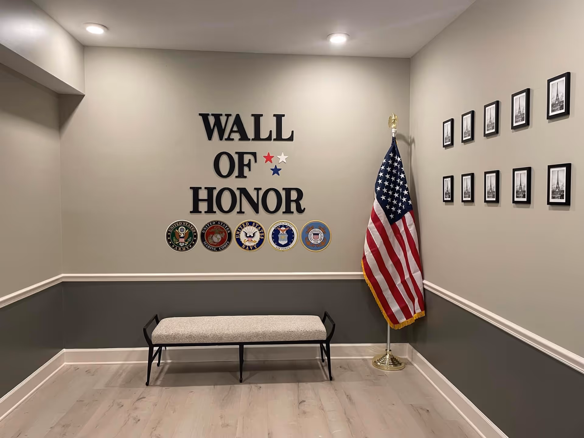A small room with a bench in front of a wall displaying the words 'WALL OF HONOR' along with five military service emblems and three stars in red, white, and blue. An American flag stands in the corner, and a series of framed black and white photos are arranged on the adjacent wall.