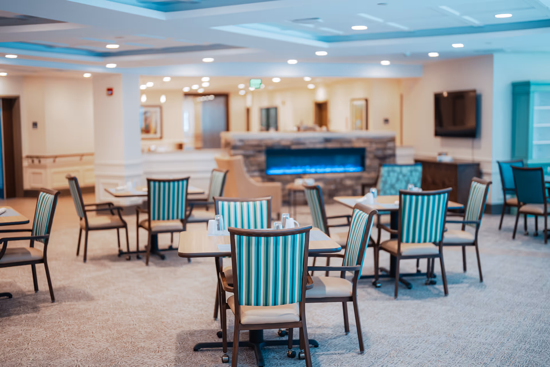 Communal dining room with multiple tables and teal-striped chairs facing a stone fireplace and wall-mounted TV.