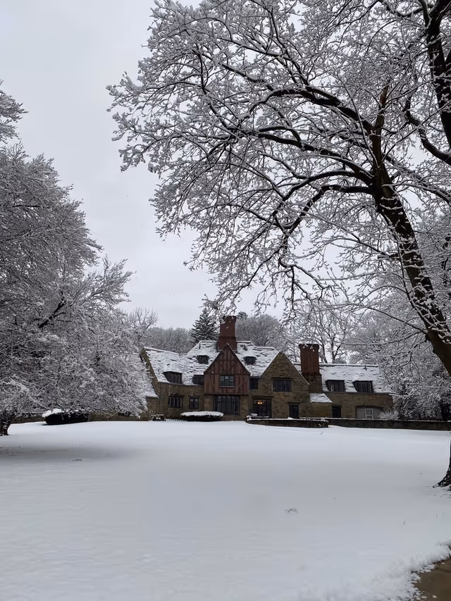 A large stone building with multiple chimneys and a steep roof covered in snow, surrounded by snow-covered trees and a snow-blanketed lawn under a gray sky.