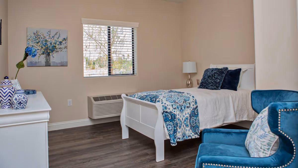 A neatly arranged bedroom with a white single bed featuring a blue and white patterned blanket and dark blue pillows. There is a blue upholstered armchair with a floral cushion in the foreground. A white dresser with decorative blue and white vases is on the left side, and a window with blinds is centered on the back wall. A table lamp is placed on a small nightstand next to the bed. The room has beige walls and dark wood flooring.