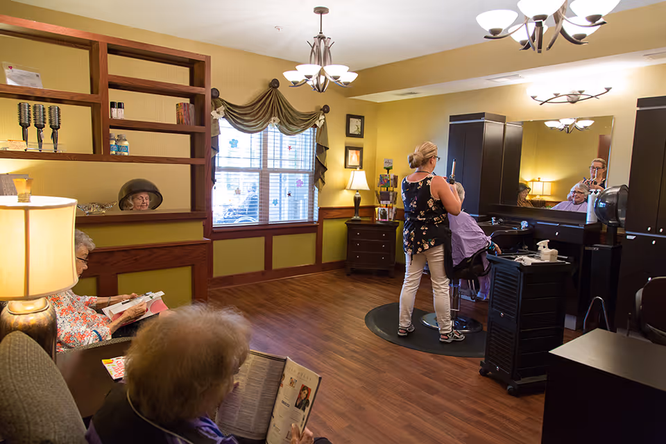 A senior living facility salon area with a hairdresser styling an elderly woman's hair. Two other elderly women are seated nearby, reading magazines. The room has wooden floors, yellow walls, a large window with drapes, and salon equipment including a hair dryer and mirrors.