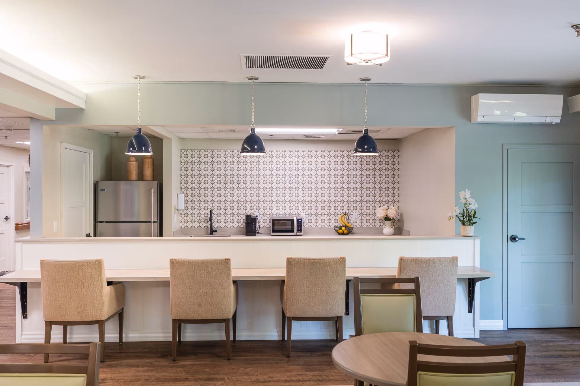 A modern kitchen area with a long counter and four beige upholstered chairs. The kitchen features a patterned tile backsplash, a stainless steel refrigerator, a microwave, a coffee maker, and decorative items including a fruit basket and flowers. Three blue pendant lights hang above the counter, and there is a round table with chairs in the foreground.