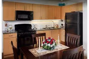 A modern kitchen and dining area featuring wooden cabinets, a black refrigerator, a black microwave, and a black stove. A wooden dining table with four chairs is set with a floral centerpiece and two lit candles on a lace table runner.
