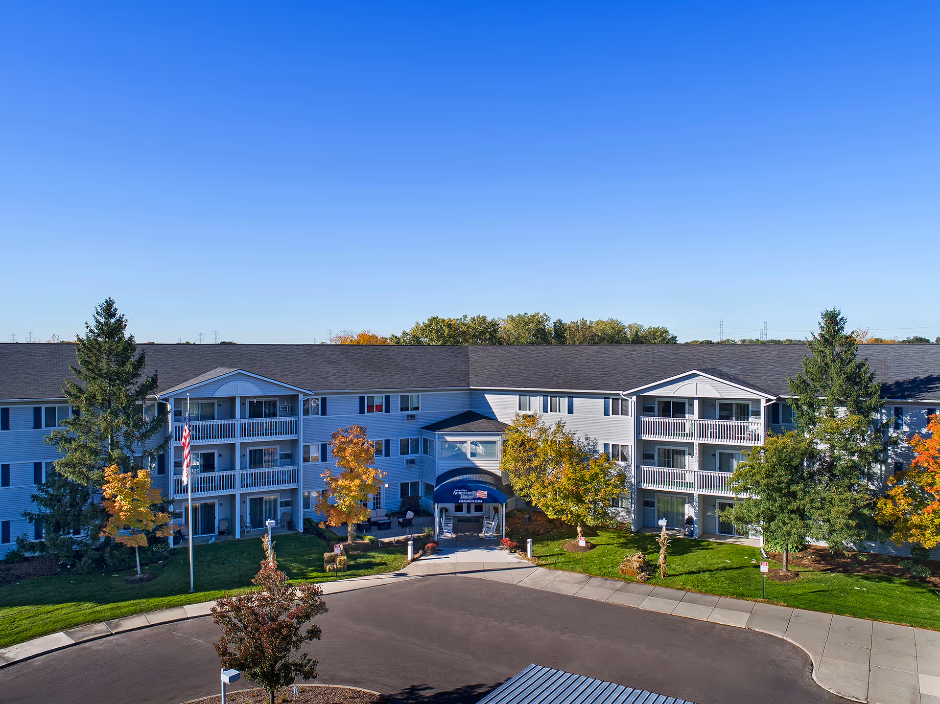 Front exterior of a three-story senior living building with balconies, an entrance canopy, landscaped trees, and an American flag.