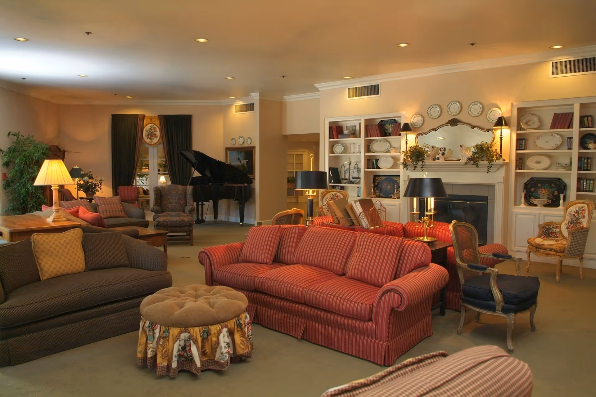 A cozy living room in an assisted living community featuring multiple upholstered sofas and chairs, a round tufted ottoman with a decorative skirt, a grand piano in the corner, built-in white shelves with decorative plates and books, a fireplace with a mirror and wall sconces above it, and warm lighting from table lamps.