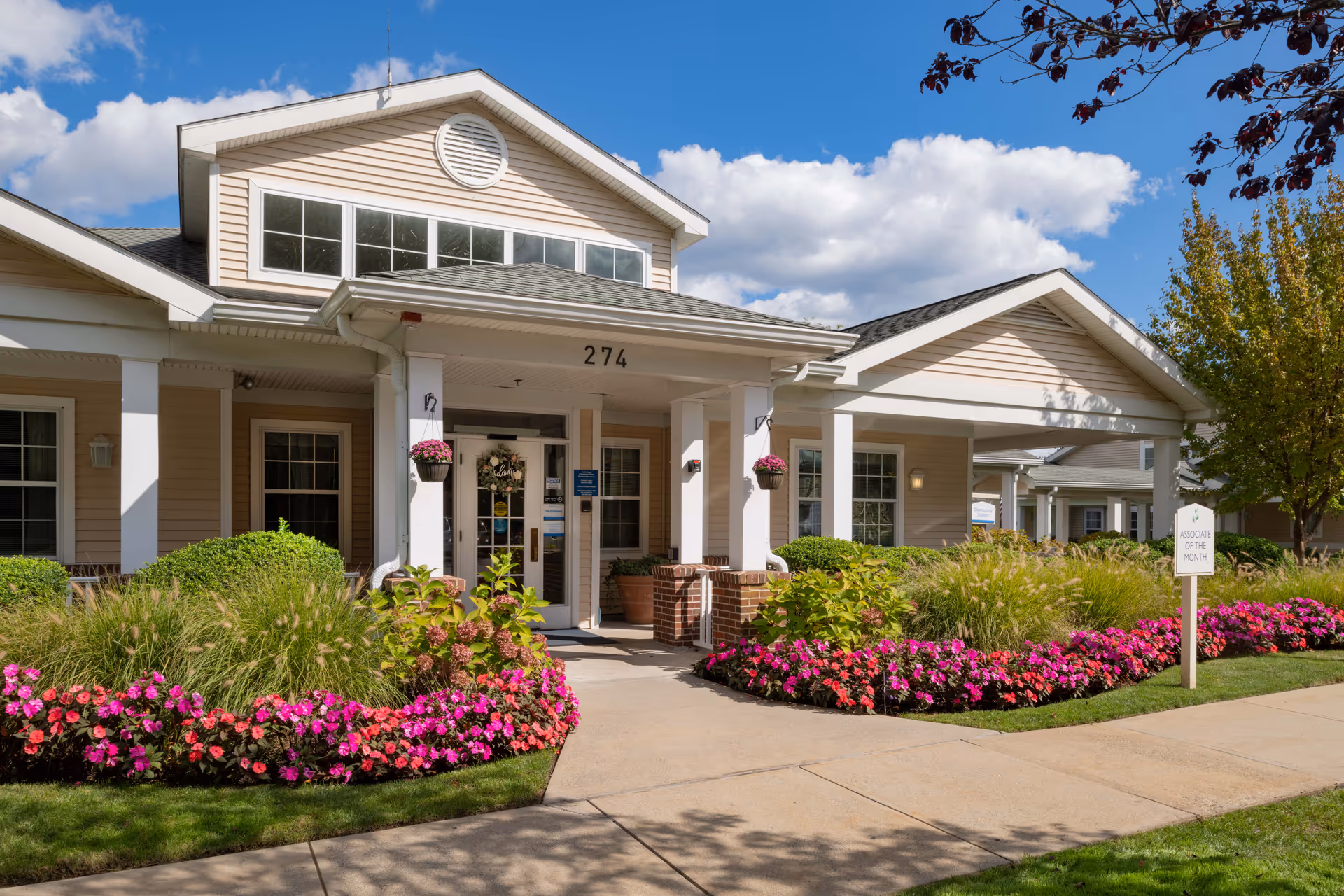 Front exterior view of The Cottages of Dartmouth Village building with beige siding, white columns, and a covered entrance. The entrance is surrounded by well-maintained landscaping with green shrubs and vibrant pink flowers. A sidewalk leads to the entrance under a partly cloudy blue sky.
