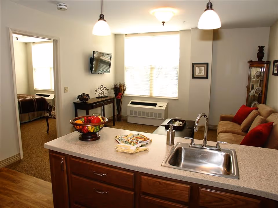 Interior view of a senior living facility apartment showing a kitchen counter with a sink and a fruit bowl, a living room area with a beige couch and red pillows, a small table with decorative items, a wall-mounted TV, and a bedroom visible through an open doorway.