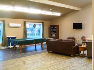 A spacious common room with a pool table covered in green felt, a brown leather couch, a wall-mounted flat screen TV, a bookshelf filled with books, and large windows with green curtains letting in natural light. The room has wood flooring and beige walls.