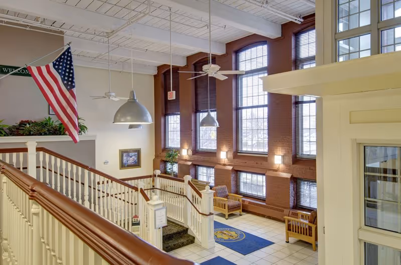 Interior view of a senior living facility with a staircase featuring white railings and wooden handrails. Large windows with brown frames allow natural light to fill the space. There are ceiling fans and hanging lights above, an American flag hanging on the left side, and wooden chairs placed near the windows. The floor is tiled with blue mats displaying a logo.