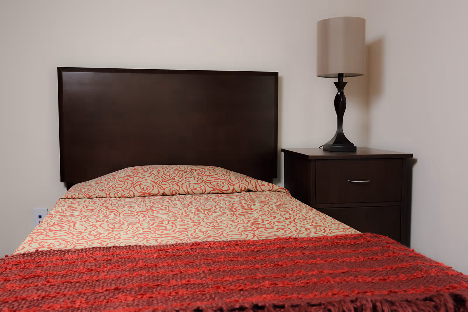 A single bed with a dark wooden headboard, a beige bedspread with a red circular pattern, and a red knitted blanket at the foot of the bed. Next to the bed is a dark wooden nightstand with a drawer and a modern table lamp with a beige lampshade.