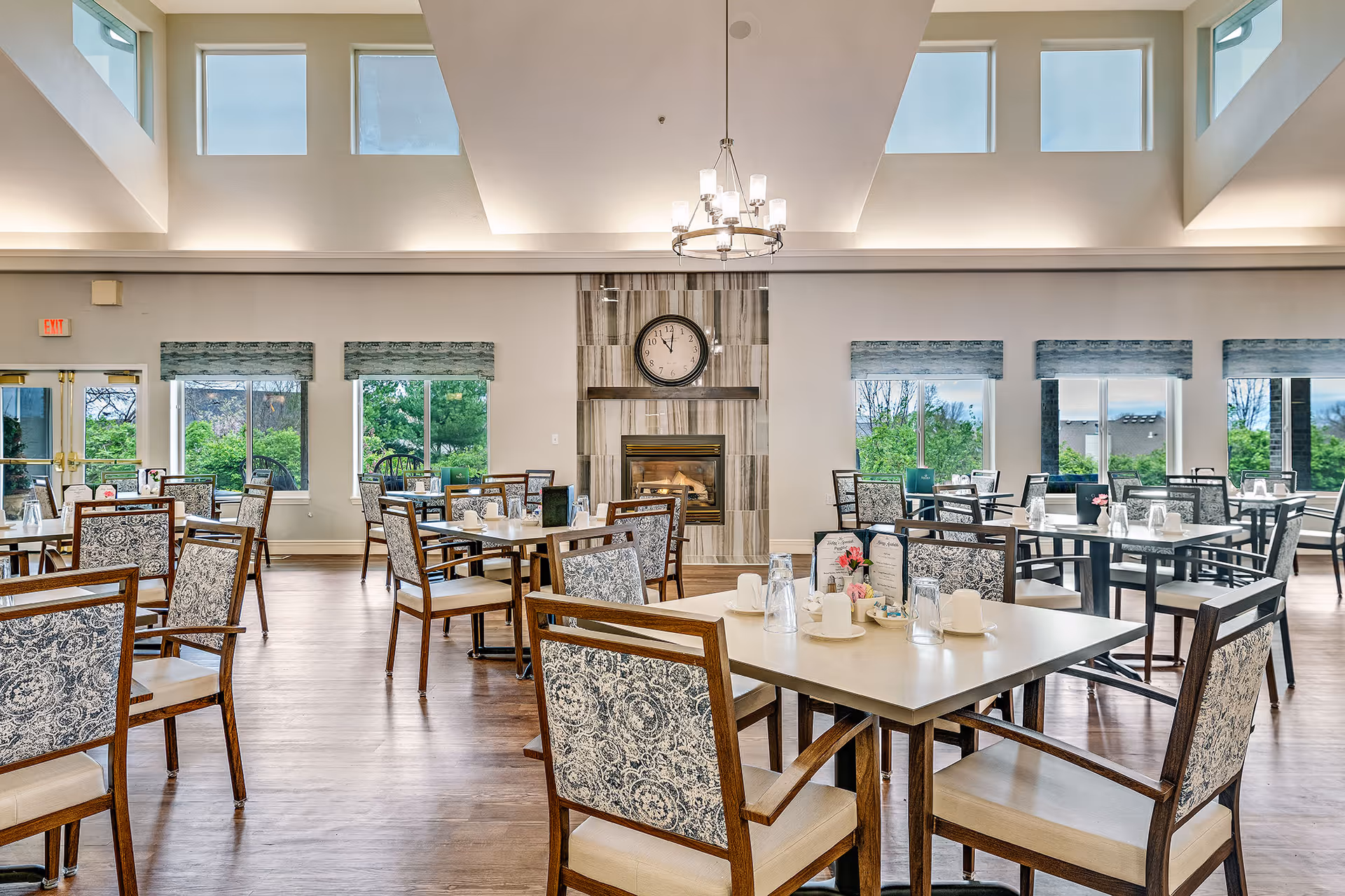 A bright and spacious dining room with multiple tables and chairs arranged neatly. The chairs have patterned upholstery and the tables are set with cups, glasses, and menus. There is a fireplace with a clock above it on the far wall, and large windows letting in natural light with views of greenery outside. A chandelier hangs from the ceiling, and the room has a warm, inviting atmosphere.