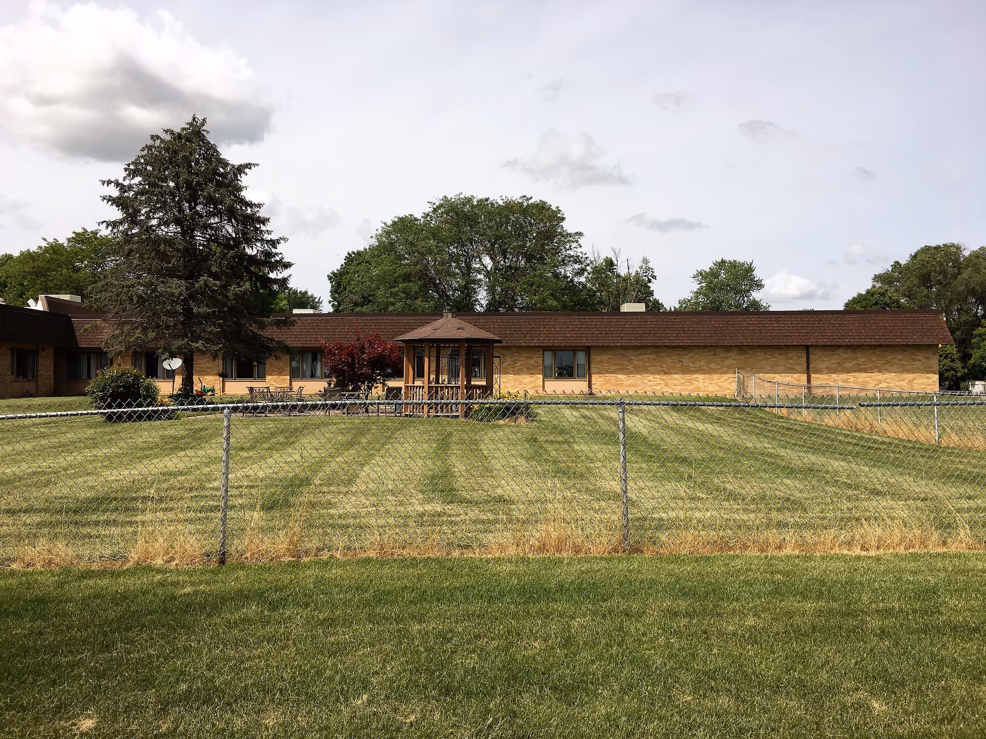 Single-story brick building with a fenced lawn, a small gazebo and trees under a cloudy sky.