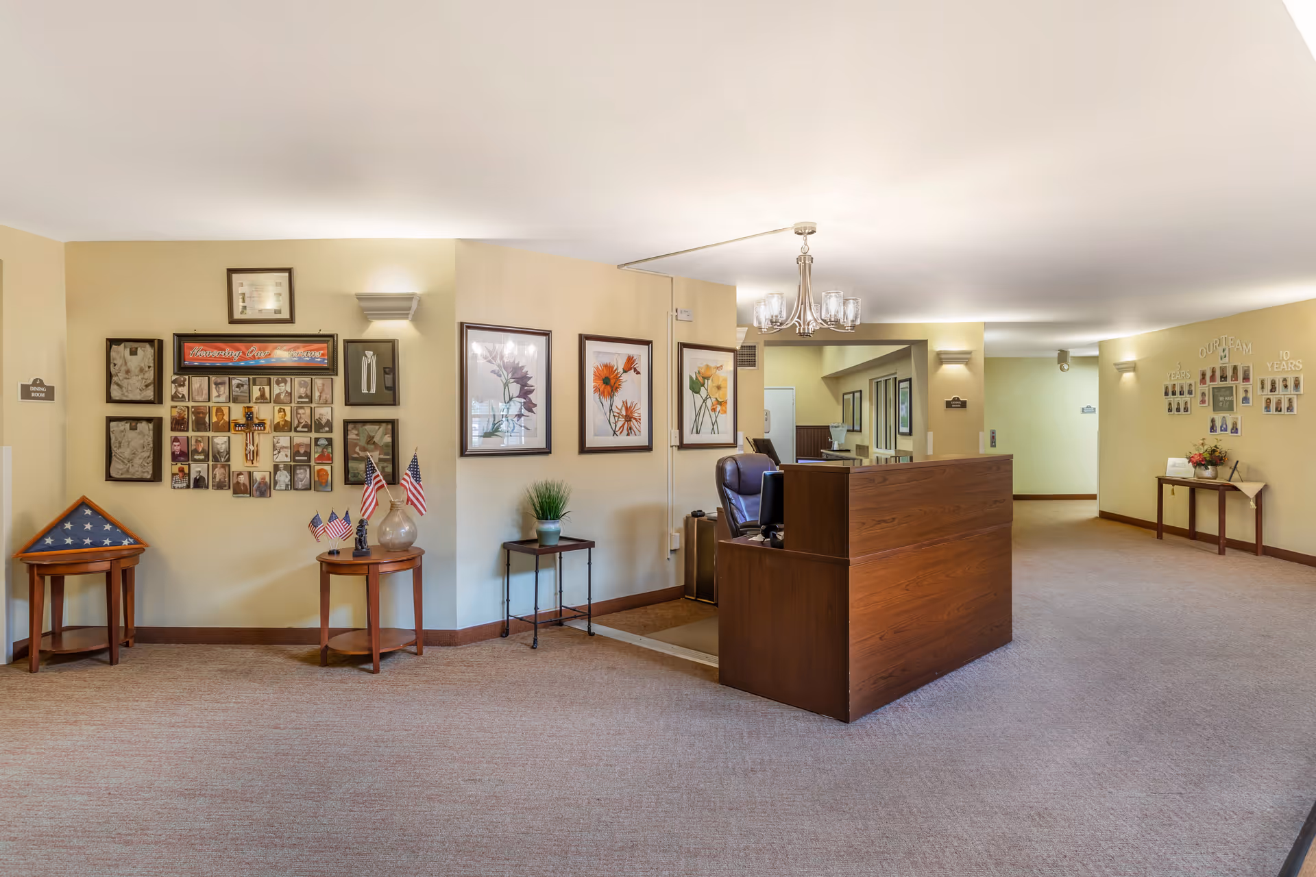 Reception area in a senior living facility with a wooden front desk, a chair behind it, and walls decorated with framed pictures and floral artwork. There are small tables with American flags and a folded flag display case. The carpeted floor and soft lighting create a welcoming atmosphere.