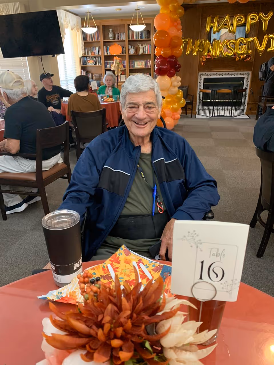 An elderly man smiling at a table with fall decorations and a "Happy Thanksgiving" balloon display in a communal dining room.