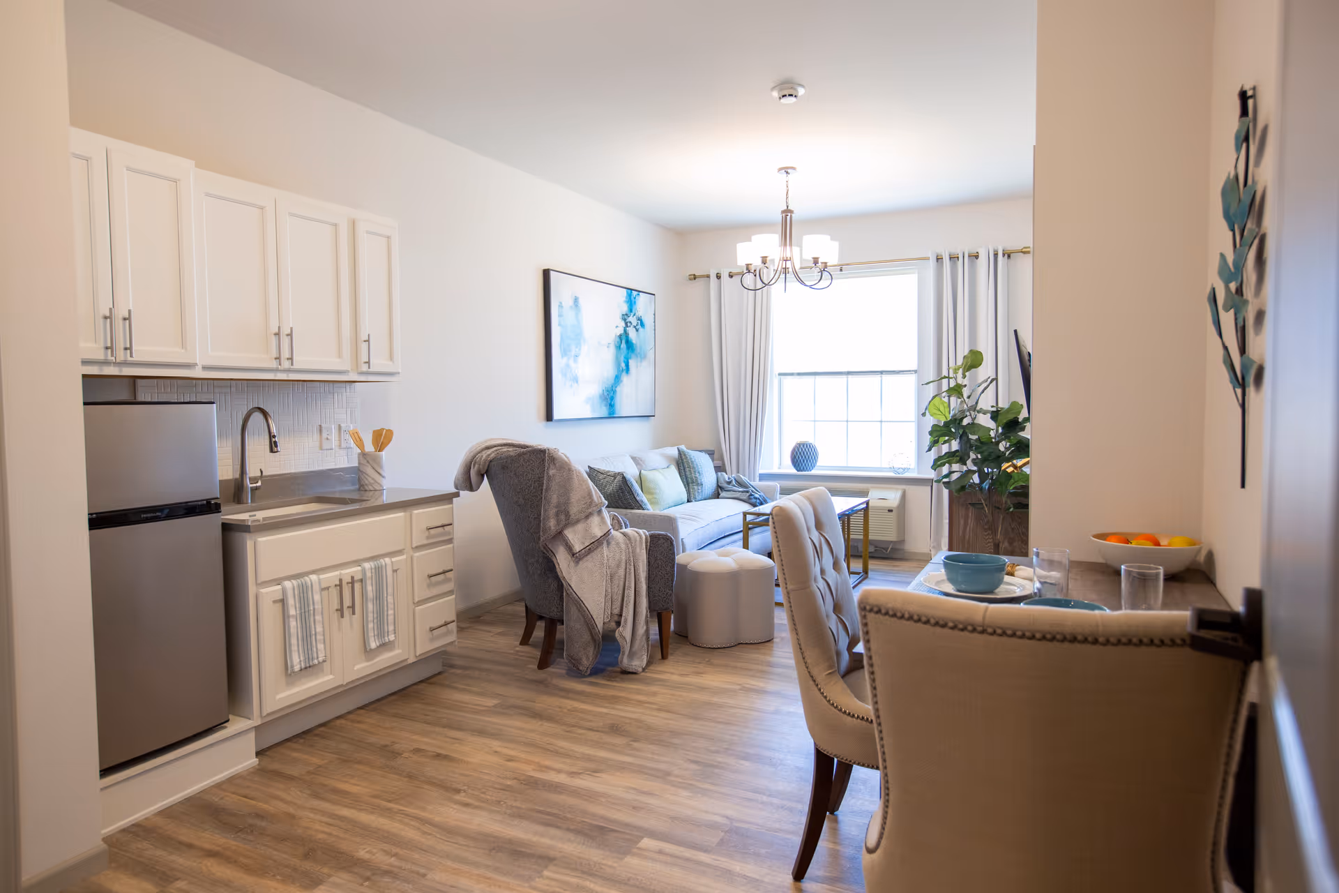 Bright furnished studio apartment interior with a small kitchenette, dining table and chairs, and a living area with a sofa and window.