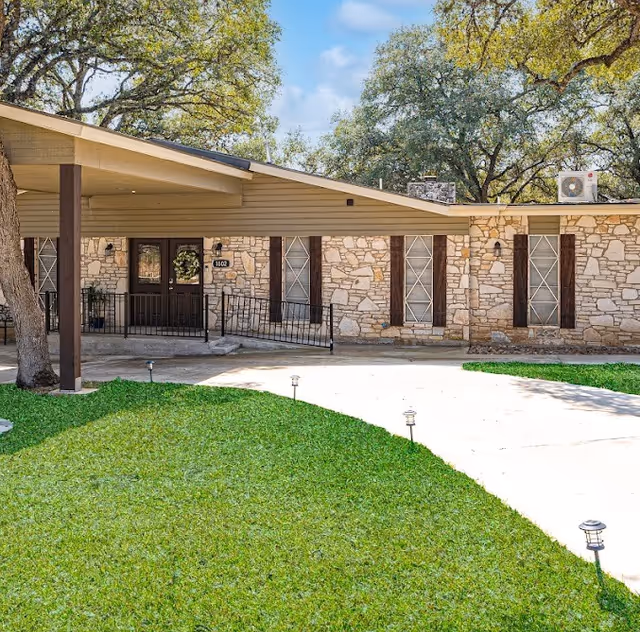 Front exterior of a single-story stone assisted living home with a covered entry, wheelchair ramp, circular driveway and grassy lawn.